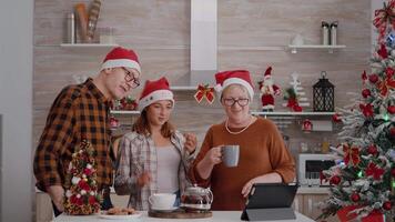 Grandparents with granddaughter greeting remote friends during online videocall meeting in xmas decorated kitchen. Happy family enjoying winter season celebrating christmas video