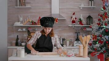 Portrait of happy child wearing apron making homemade dough using rollling pin preparing traditional delicious gingerbread dessert. Girl standing at table in xmas decorated kitchen enjoying christmas video