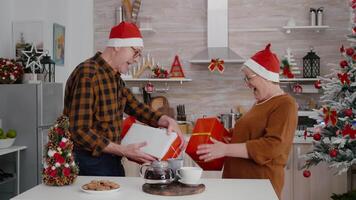 Senior couple with sanata claus hat sharing xmas wrapper gift with ribbon on it during christmas holiday in decorated kitchen. Happy family enjoying christmastime celebrating winter season video