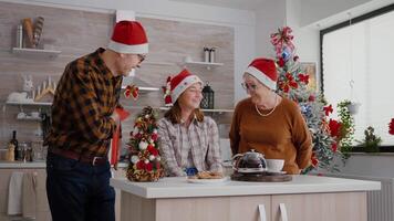 Grandparents giving wrapper xmas gift with ribbon on it to granddaughter standing at table in decorated kitchen. Happy family enjoying winter season celebrating christmas holiday video