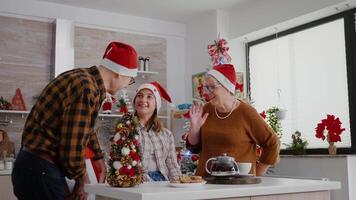 Happy family enjoying winter holiday celebrating christmas season standing at table in decorated kitchen. Grandparents surprising grandchild wrapper present with ribbon on it during christmastime video