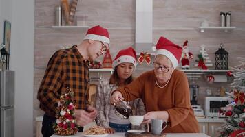 familie met de kerstman hoed te hard rijden Kerstmis vakantie samen genieten van kersttijd glimlachen in versierd keuken. grootouders drinken koffie terwijl kleinkind aan het eten heerlijk chocola gebakken koekjes video