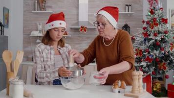 Granddaughter putting flour ingredient in strainer while granddaughter sift preparing homemade delicious cookie dessert in xmas decorated culinary kitchen. Happy family celebrating christmas holidays video