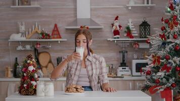 ritratto di bambini guardare in telecamera mentre seduta a tavolo nel natale decorato cucina mangiare al forno delizioso biscotti potabile latte. caucasico ragazza godendo Natale vacanza stagione video