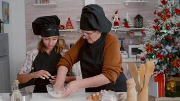 Granddaughter with grandma preparing traditional homemade dough baking traditional cookies dessert in culinary kitchen during christmastime. Family celebrating christmas holidays enjoying cooking video
