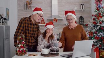 Grandparents with granddaughter greeting remote friends during online videocall meeting celebrating christmas holiday in xmas decorated culinary kitchen. Happy family enjoying winter season together video