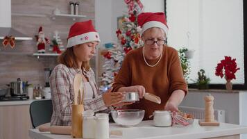 senior vrouw zetten meel in metaalachtig zeef terwijl kleindochter ziften ingrediënt in kom voorbereidingen treffen Kerstmis traditioneel koekje. familie genieten van Kerstmis vakantie samen in versierd keuken video