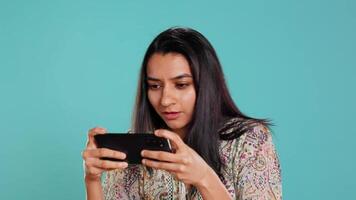 Lycklig indisk kvinna underhöll förbi Videospel på smartphone, njuter fritid tid. gamer njuter spel på telefon, har roligt besegra fiender, isolerat över studio bakgrund, kamera b video