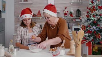Granddaughter with granddaughter breaking egg in kitchen bowl with flour ingredient preparing homemade dough. Happy family enjoying making xmas cookie delicious dessert during christamastime video