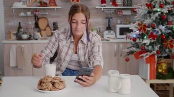 feliz menina desfrutando inverno feriado sentado às mesa dentro natal decorado cozinha navegando em Smartphone assistindo festivo usando telefone. menina a comemorar Natal estação comendo cozido biscoitos video