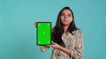 Portrait of indian woman doing influencer marketing using green screen tablet, isolated over studio background. Smiling person holding empty copy space mockup device, camera A video