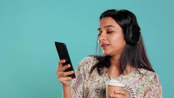 Joyous woman listening music, singing lyrics from phone screen, enjoying beverage. Cheerful person wearing headphones, listening songs, head bopping and holding coffee cup, studio background, camera B video