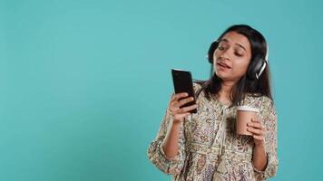 Joyous woman listening music, having fun, dancing on rhythm, enjoying beverage. Upbeat indian person wearing headphones, doing funky movements, holding coffee cup, studio background, camera A video