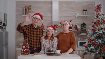 pov de contento familia vistiendo Papa Noel sombrero saludo remoto amigos durante en línea videollamada conferencia reunión en pie a mesa en Navidad decorado cocina. abuelos disfrutando Navidad fiesta video