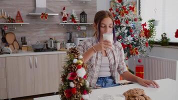 Child sitting at table in decorated kitchen eating traditional xmas baked cookies drinking milk enjoying winter season. Girl celebrating christmas holiday during morning at home video