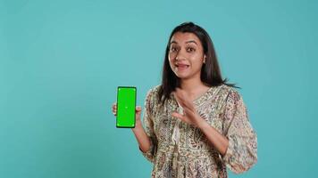 kvinna presenter grön skärm mobil telefon, isolerat över studio bakgrund. glad indisk person innehav kopia Plats krom nyckel smartphone Begagnade för reklam märken, kamera en video