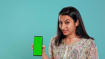 Tech content creator doing influencer marketing using green screen smartphone, isolated over studio background. Indian woman holding empty copy space mockup phone, doing review, camera B video