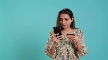 indisk kvinna upphetsad till köp av grejer, tillsats betalning metod på mobil telefon hemsida, isolerat över studio bakgrund. extatisk klient gör internet handla, skriver debitera kort info, kamera en video