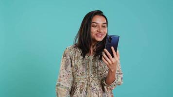 Smiling woman having friendly conversations during teleconference meeting using smartphone, studio background. Indian person having fun catching up with mates during online videocall, camera A video