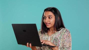 Indian woman typing on laptop keyboard and cursing, feeling angry at work. Person writing emails on notebook, complaining loudly, isolated over studio background, camera B video