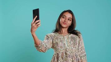 alegre indiano mulher usando Smartphone para levar selfies e postar eles em social meios de comunicação. feliz pessoa levando fotos usando telefone selfie Câmera, isolado sobre estúdio fundo, Câmera uma video