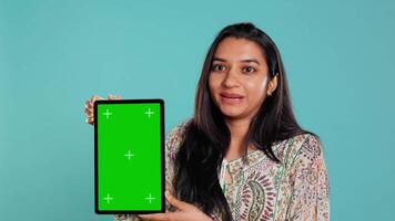 porträtt av indisk kvinna håller på med influencer marknadsföring använder sig av grön skärm läsplatta, isolerat över studio bakgrund. leende person innehav tömma kopia Plats attrapp enhet, kamera b video