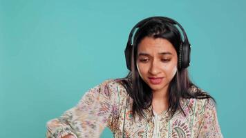 Indian woman playing music, having fun moving on rhythm, pretending to use turntables. Upbeat DJ wearing headphones, producing beats, isolated over studio background, camera B video