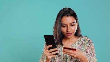 Indian woman excited to purchasing stuff, adding payment method on mobile phone website, isolated over studio background. Ecstatic client does internet shopping, typing debit card info, camera B video