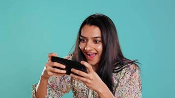 Happy woman playing videogames on smartphone, celebrating after defeating foes, doing clenching hand gesture. Player swiping phone screen, thrilled about winning game, studio background, camera B video