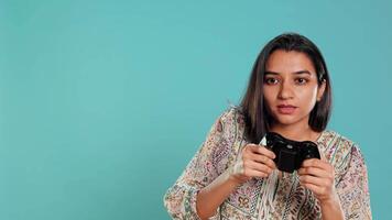 Cheerful woman playing videogames with controller, studio background. Indian gamer smiling, having fun by participating in PvP online multiplayer game using console system, camera B video