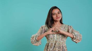 portret van vrolijk vriendelijk Indisch vrouw aan het doen hart symbool vorm gebaar met handen. vrolijk koesteren persoon tonen liefde gebaren, geïsoleerd over- studio achtergrond, camera een video