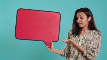 Smiling indian woman holding red speech bubble sign of empty copy space for message. Joyous person presenting thought bubble cardboard used as promotion concept, studio background, camera A video