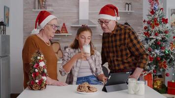Happy family watching online xmas movie on tablet computer enjoying holiday season standing at table in decorated kitchen. Grandchild eating baked cookies celebrating christmas tradition video
