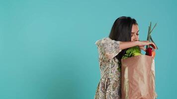 Indien femme avec réutilisable papier sac dans mains à la recherche à pomme, vivant en bonne santé mode de vie. écologie amoureux en portant achats sac avec Naturel produire, studio arrière-plan, caméra une video