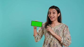 Smiling woman showing isolated screen mobile phone, doing promotion, studio background. Upbeat indian person holding copy space mockup cellphone used for advertising brands, camera A video