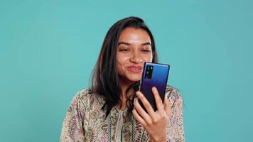 Smiling woman having friendly conversations during teleconference meeting using smartphone, studio background. Indian person having fun catching up with mates during online videocall, camera B video