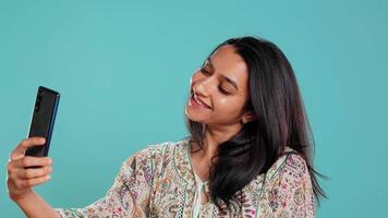 alegre indio mujer sonriente para autofotos, destino ellos en social medios de comunicación utilizando Teléfono móvil. jubiloso niña tomando imágenes utilizando teléfono selfie cámara, aislado terminado estudio fondo, cámara si video