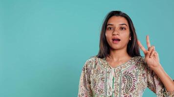 Jolly indian person doing peace victory hand sign gesture, studio backdrop. Portrait of upbeat woman, feeling confident, celebrating win, isolated over studio background, camera B video