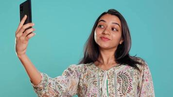 vrolijk Indisch vrouw gebruik makend van smartphone naar nemen selfies en post hen Aan sociaal media. gelukkig persoon nemen foto's gebruik makend van telefoon selfie camera, geïsoleerd over- studio achtergrond, camera b video