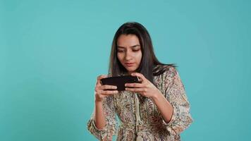 Lycklig indisk kvinna underhöll förbi Videospel på smartphone, njuter fritid tid. gamer njuter spel på telefon, har roligt besegra fiender, isolerat över studio bakgrund, kamera en video