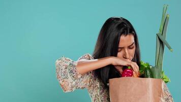 Woman with paper bag with vegetables testing quality, looking at cucumber, isolated over studio background. Person inspecting groceries after buying them from zero waste shop, camera B video