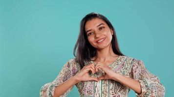 portret van vrolijk vriendelijk Indisch vrouw aan het doen hart symbool vorm gebaar met handen. vrolijk koesteren persoon tonen liefde gebaren, geïsoleerd over- studio achtergrond, camera b video