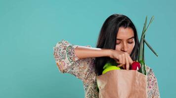 Woman with recycled paper bag in hands filled with vegetables testing quality, studio background. Sustainable living person inspecting groceries after doing purchases in shop, camera B video