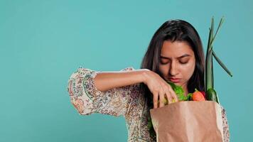 Indian woman with paper bag with vegetables and fruits testing quality, looking at orange, studio background. Vegetarian verifying groceries are ripe after buying them from zero waste shop, camera B video