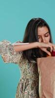 Vertical Woman with paper bag with vegetables testing quality, looking at cucumber, isolated over studio background. Person inspecting groceries after buying them from zero waste shop, camera A video