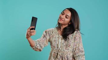 alegre indio mujer sonriente para autofotos, destino ellos en social medios de comunicación utilizando Teléfono móvil. jubiloso niña tomando imágenes utilizando teléfono selfie cámara, aislado terminado estudio fondo, cámara un video