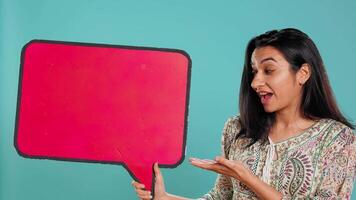 Smiling indian woman holding red speech bubble sign of empty copy space for message. Joyous person presenting thought bubble cardboard used as promotion concept, studio background, camera B video