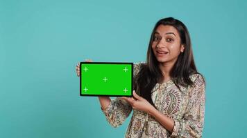 Happy woman presenting green screen tablet, isolated over studio background. Joyous indian person holding copy space chroma key device used for advertising brands, camera A video