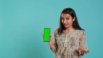 Tech content creator doing influencer marketing using green screen smartphone, isolated over studio background. Indian woman holding empty copy space mockup phone, doing review, camera A video