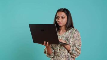 indiano donna digitando su il computer portatile tastiera e imprecando, sensazione arrabbiato a opera. persona scrittura messaggi di posta elettronica su taccuino, lamentarsi a voce alta, isolato al di sopra di studio sfondo, telecamera un' video